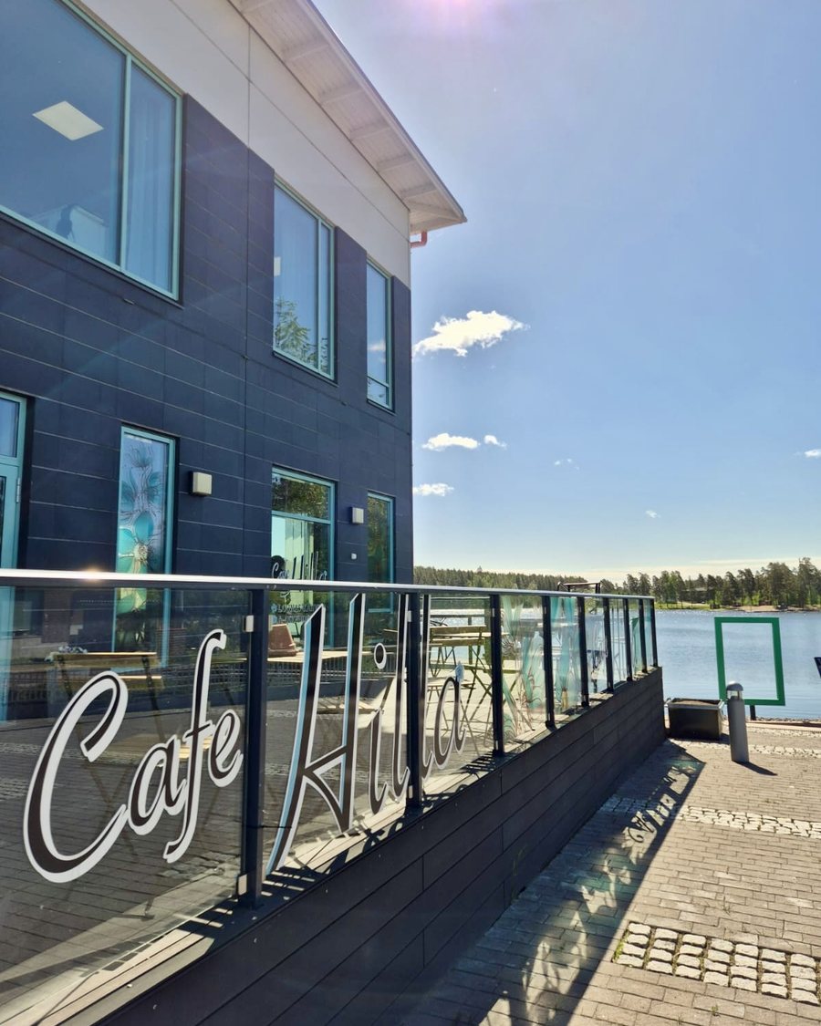 Cafe Hillan lasikaide ja terassi, jossa lukee Cafe Hilla, taustalla Huutjärvi
